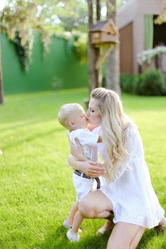 Young happy mother playing and sitting with little kid on grass in yard and kissing son. Concept of motherhood and child.