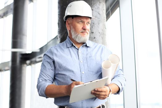 Confident mature business man in hardhat holding blueprint and looking away while standing indoors