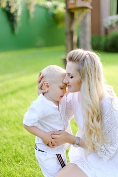 Young pretty mother playing and sitting with little kid on grass in yard and kissing son. Concept of motherhood and child.