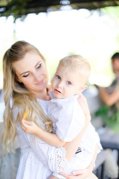 Young blonde mother holding baby outside and wearing white clothes. Concept of motherhood and child.