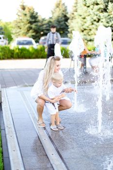 Young happy mother sitting with little baby outside near fountain. Concept of walking in city and motherhood.