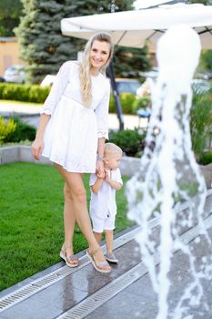 Little baby with young mother standing on grass near fountain. Concept of childhood and summer.