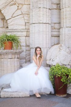 Caucasian charming fiancee sitting near ancient columns and wearing white dress. Concept of bridal photo session and wedding.