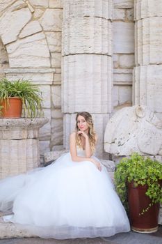 Caucasian nice fiancee sitting near ancient columns and wearing white dress. Concept of bridal photo session and wedding.