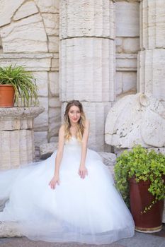 Caucasian gorgeous fiancee sitting near ancient columns and wearing white dress. Concept of bridal photo session and wedding.