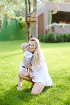 Young happy mother playing and sitting with little kid on grass in yard. Concept of motherhood and child.