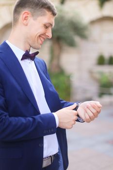 Happy smiling groom waiting for bridea and looking at watch, wearing dark blue suit. Concept of bridal photo session and wedding.