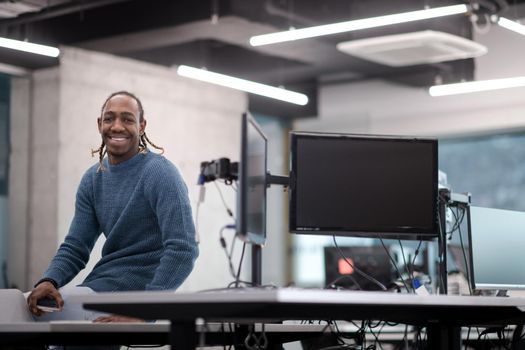 Portrait of young smiling african american male software developer sitting on office desk at modern startup office