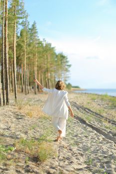 Back view of female person walking on sand beach and wearing white clothes. Concept of summer vacations.