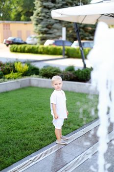 Little child standing on grass near fountain and wearing white clothes. Concept of childhood and summer.