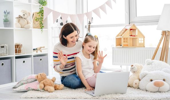 Cute little girl with pretty mother doing video calling on laptop in cozy children room