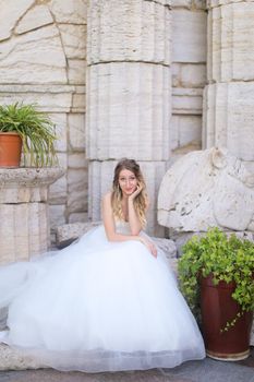 Caucasian pretty fiancee sitting near ancient columns and wearing white dress. Concept of bridal photo session and wedding.