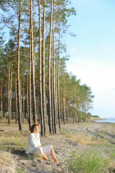 Young female person sitting on sand beach with trees in backgroundand wearing white clothes. Concept of freedom and summer vacations