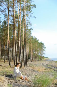 Young lady sitting on sand beach with trees in backgroundand wearing white clothes. Concept of freedom and summer vacations