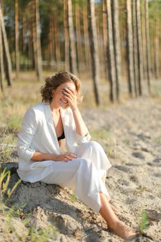 Young pretty woman sitting on sand beach with trees in backgroundand wearing white clothes. Concept of freedom and summer vacations