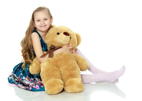 Beautiful little girl with long blond hair in a smart blue dress. The girl hugs a large teddy bear.