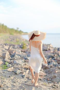 Back view of european woman in hat with red ribbon walking on shingle beach. Concept of summer vacations and resort.