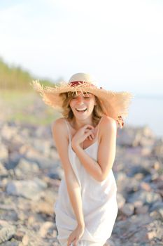 Young smiling woman in hat with red ribbon standing on shingle beach. Concept of summer vacations and resort.