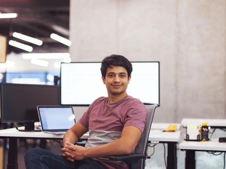 Portrait of young indian successful male software developer sitting on desk at modern startup office