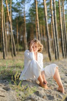 Young blonde woman sitting on sand beach with trees in backgroundand wearing white clothes. Concept of freedom and summer vacations