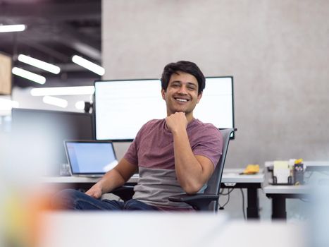 Portrait of young indian successful male software developer sitting on desk at modern startup office