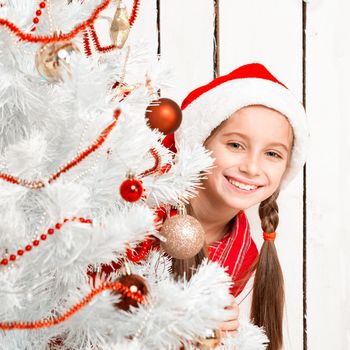 smiling little girl in red hat peeking out from behind a decorated new year tree