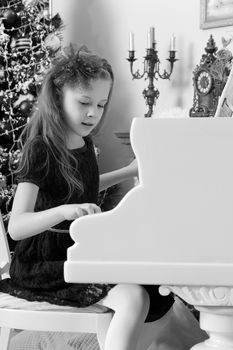 Beautiful, elegant little girl holding hands on the keys of a white Grand piano. Girl playing at the Christmas concert in the music school.Black-and-white photo. Retro style.