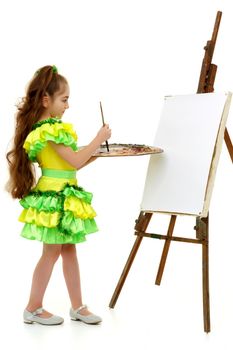 Cute little girl with a palette and brush near the easel. At the easel a blank white sheet of paper on which you can draw a picture or apply any advertising inscription. The concept of art, children's creativity.