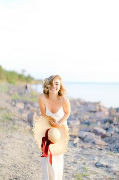 Caucasian cute girl walking on rocky beach with hant in hands and wearing dress. Concept of summer vacations and fashion.