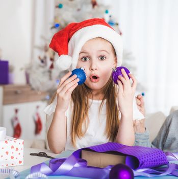 Surprised little girl in santa hat holding blue and purple balls at Christmas
