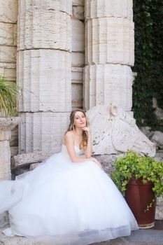 Caucasian fiancee sitting near ancient columns and wearing white dress. Concept of bridal photo session and wedding.