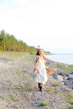 Caucasian blonde girl walking on shingle beach and wearing summer coat. Concept of seasonal fashion and summer vacations.