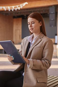 young caucasian woman in coat using tablet outdoors on sunny day, reading ebook, watching movie. attractive female street portrait at fall or spring. autumn lifestyle, modern communication technology