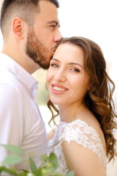 Happy caucasian groom kissing bride keeping bouquet of flowers. Concept of love and wedding photo session.