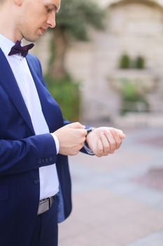 Happy handsome groom waiting for bridea and looking at watch, wearing dark blue suit. Concept of bridal photo session and wedding.