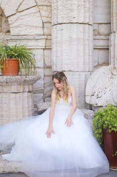 Caucasian american fiancee sitting near ancient columns and wearing white dress. Concept of bridal photo session and wedding.