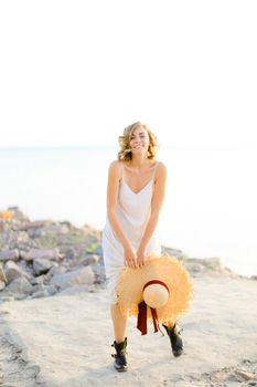 Young blonde girl walking on rocky beach with hant in hands and wearing dress. Concept of summer vacations and fashion.