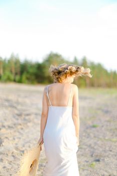 Back view of blonde girl walking on rocky beach with hant in hands and wearing dress. Concept of summer vacations and fashion.