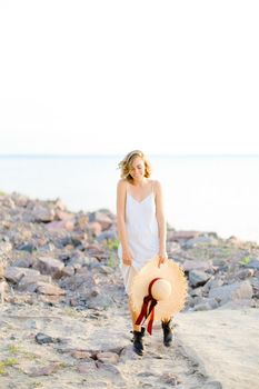 Caucasian beautiful girl walking on rocky beach with hant in hands and wearing dress. Concept of summer vacations and fashion.