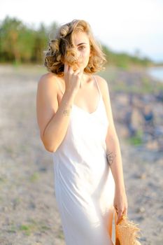 Caucasian cute blonde girl walking on rocky beach with hant in hands and wearing dress. Concept of summer vacations and fashion.