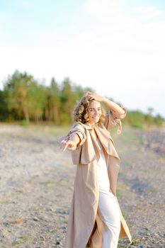 Caucasian blonde girl with outstretched hand on shingle beach wearing summer coat. Concept of seasonal fashion and summer vacations.