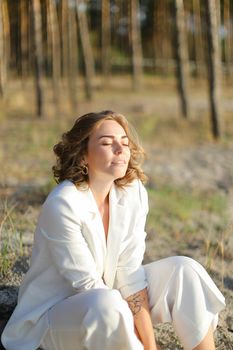 Young nice woman sitting on sand beach with trees in backgroundand wearing white clothes. Concept of freedom and summer vacations