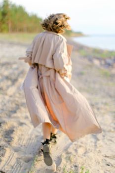 Back view of caucasian young girl walking on shingle beach and wearing summer coat. Concept of seasonal fashion and summer vacations.