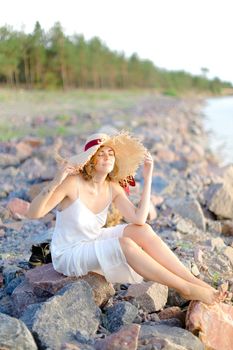 Caucasian girl in hat sitting on shingle beach. Concept of summer vacations and beauty.