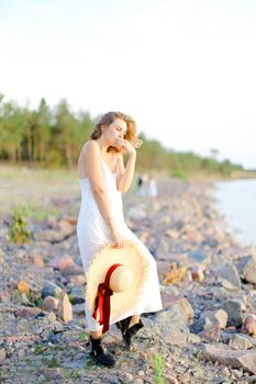 Caucasian pretty girl walking on shingle beach with hant in hands and wearing dress. Concept of summer vacations and fashion.