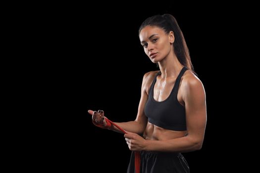 Beautiful and fit female fighter getting prepared for the fight or training, wrapping her hands with bandage tape against dark background