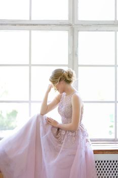 Blonde caucasian bride wearing dress and standing near window and dreaming. Concept of bridal photo session and nice fiancee.