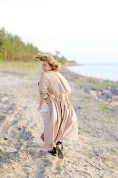 Back view of caucasian young lady walking on shingle beach and wearing summer coat. Concept of seasonal fashion and summer vacations.