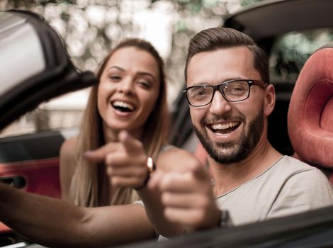 happy couple sitting inside a convertible and pointing at you.the concept of the relationship