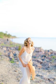 Caucasian smiling girl walking on rocky beach with hant in hands and wearing dress. Concept of summer vacations and fashion.